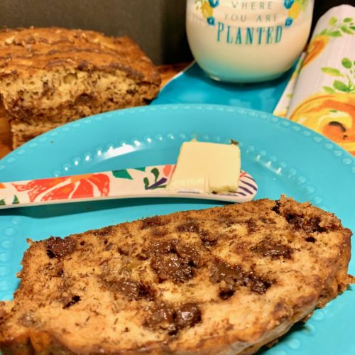 antioxidant dark chocolate chip almond banana bread on blue plate next to butter on a flower designed butter knife and a cup of milk in the background
