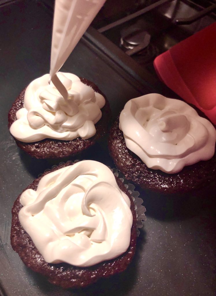 three hot cocoa cupcakes being frosted with marshmallow frosting through a pastry bag