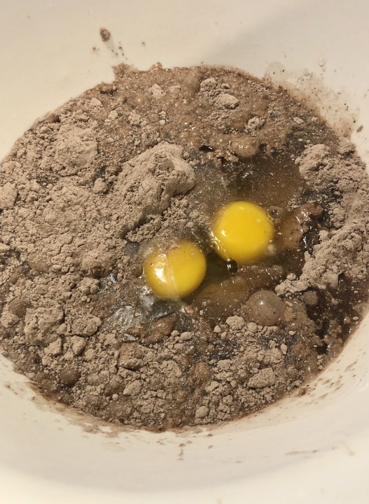 preparing boxed cake mix in a large white mixing bowl. There is a box of chocolate cake mix, vegetable oil, water and 3 eggs