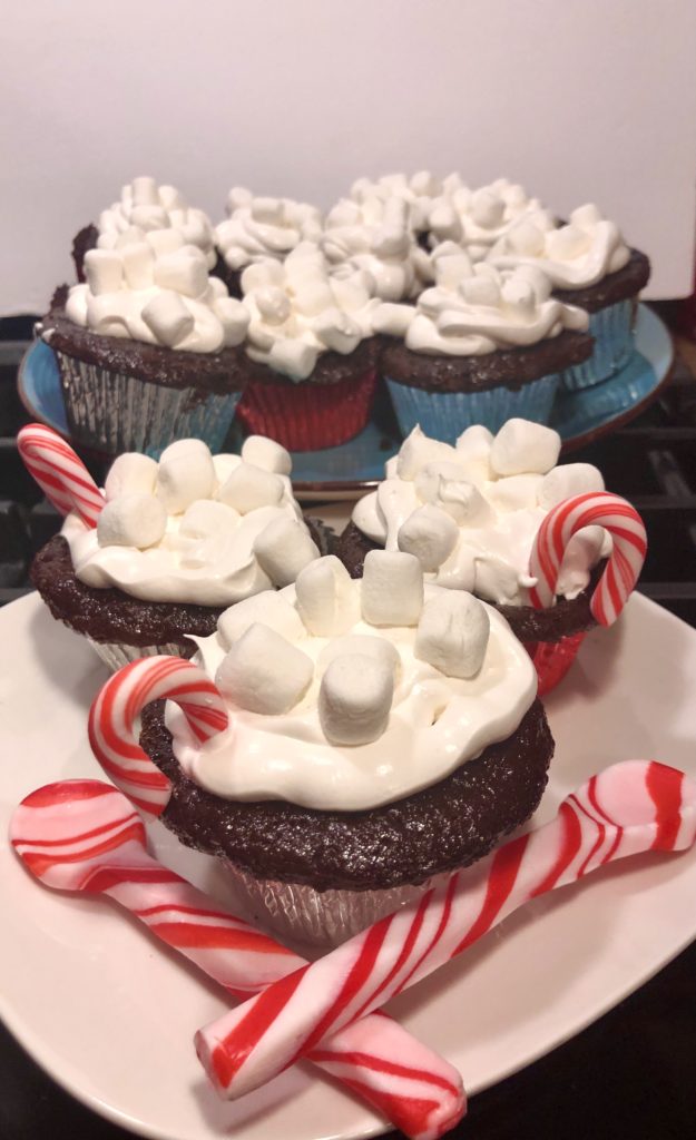 three hot cocoa cupcakes with marshmallow frosting and covered with mini marshmallows and a mini candy cane coming out from the top. Three cupcakes are pictured on a square white plate with two peppermint red and white striped spoons criss crossed. There is a blue plated pictured in the background full of decorated cupcakes.