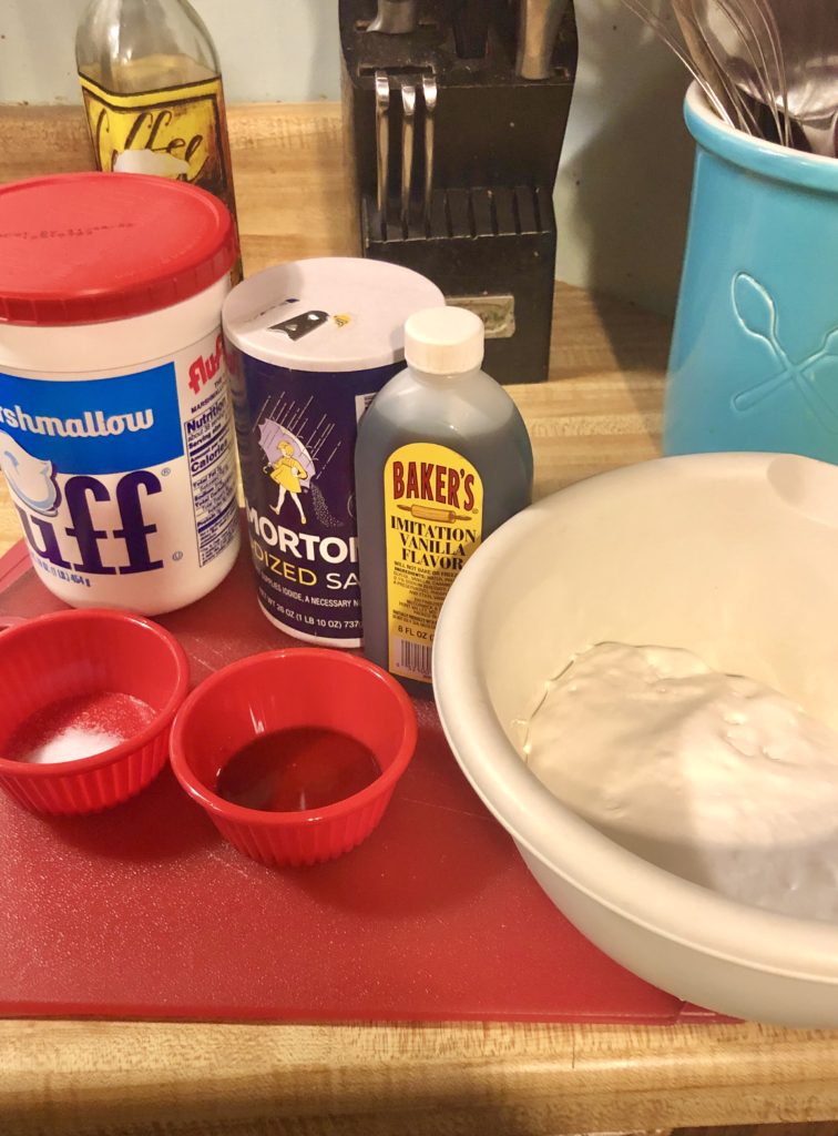 fluff, salt, vanilla extract beside fluff in white mixing bowl. There are two red ramikens with salt in one and vanilla extract in the other