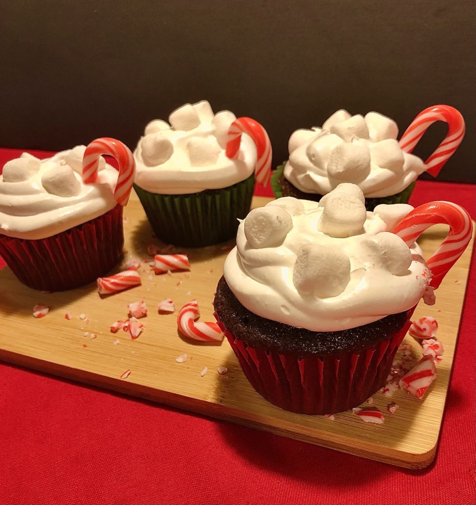 Chocolate hot cocoa cupcakes with marshmallow frosting. topped with mini marshmallows and mini candy canes to resembles a hot cup of cocoa. Four cupcakes are photographed on an oak cutting board with red fabric table and chopped up pieces of candy cane surrounding them.