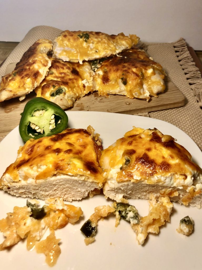 jalapeno popper smothered chicken breast with cream cheese and cheddar cheese. chicken breast is sliced in half on white plate with cutting board full of jalapeno popper smothered chicken breast in the background