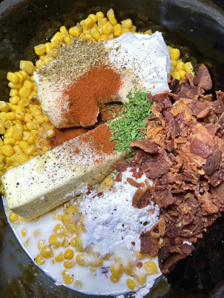 Slow cooker with corn, butter, cream cheese, bacon, cream, salt, black pepper, garlic powder, paprika and dried parsley.