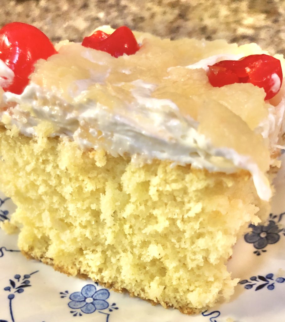 up close picture of yellow cake slice with cream cheese and cool whip frosting covered with pineapples and cherries. The cake is on a white plate with royal blue floral pattern