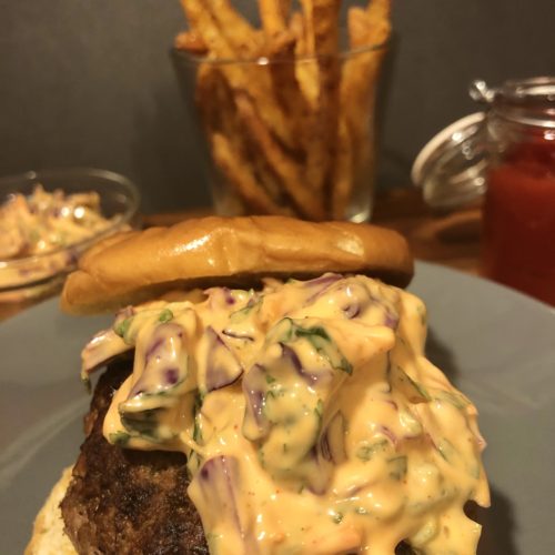 Korean burger topped with gochujang slaw between buttered hamburger buns on gray plate. In the background is a glass ramiken with additional slow, a cup with French fries and a jar of ketchup.