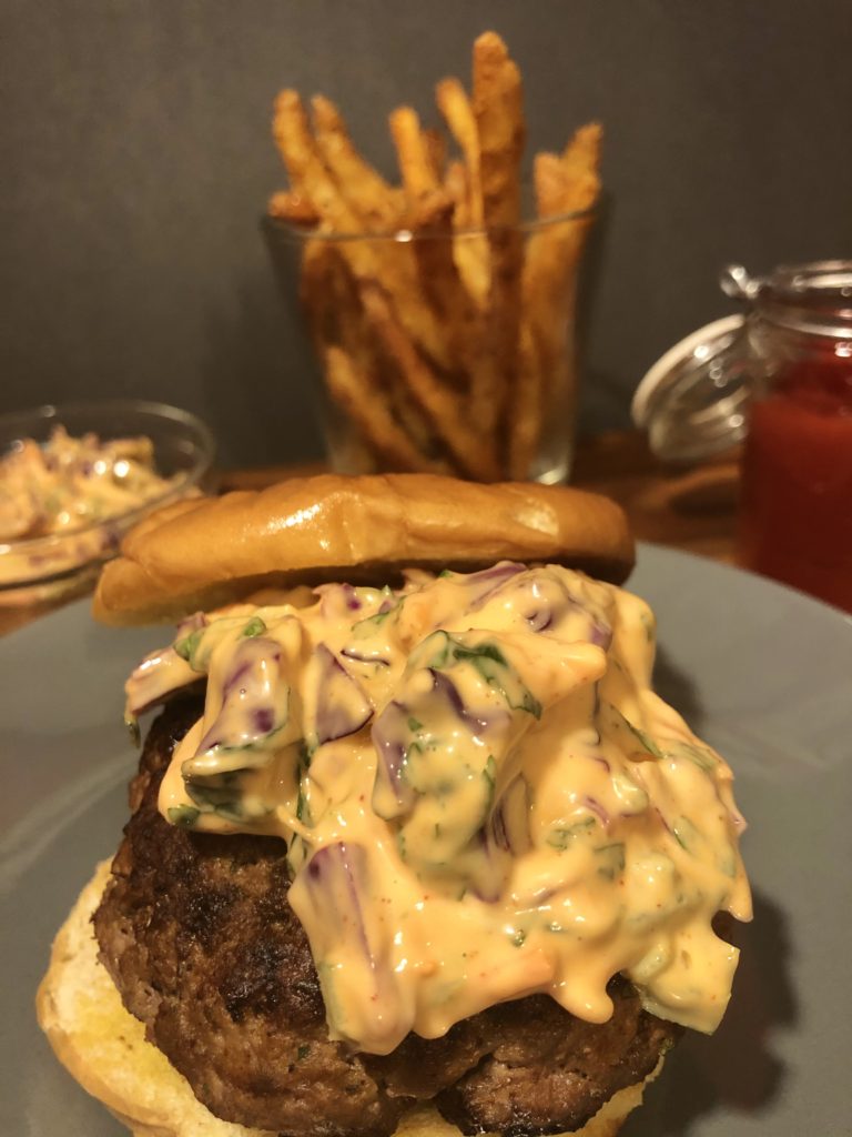 Korean burger topped with gochujang slaw between buttered hamburger buns on gray plate. In the background is a glass ramiken with additional slow, a cup with French fries and a jar of ketchup.