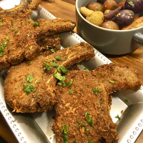 parmesan crusted pork chops on long silver plate on wooden cutting board with gray towel and gray bowl of roasted baby potatoes and rainbow carrots in the background