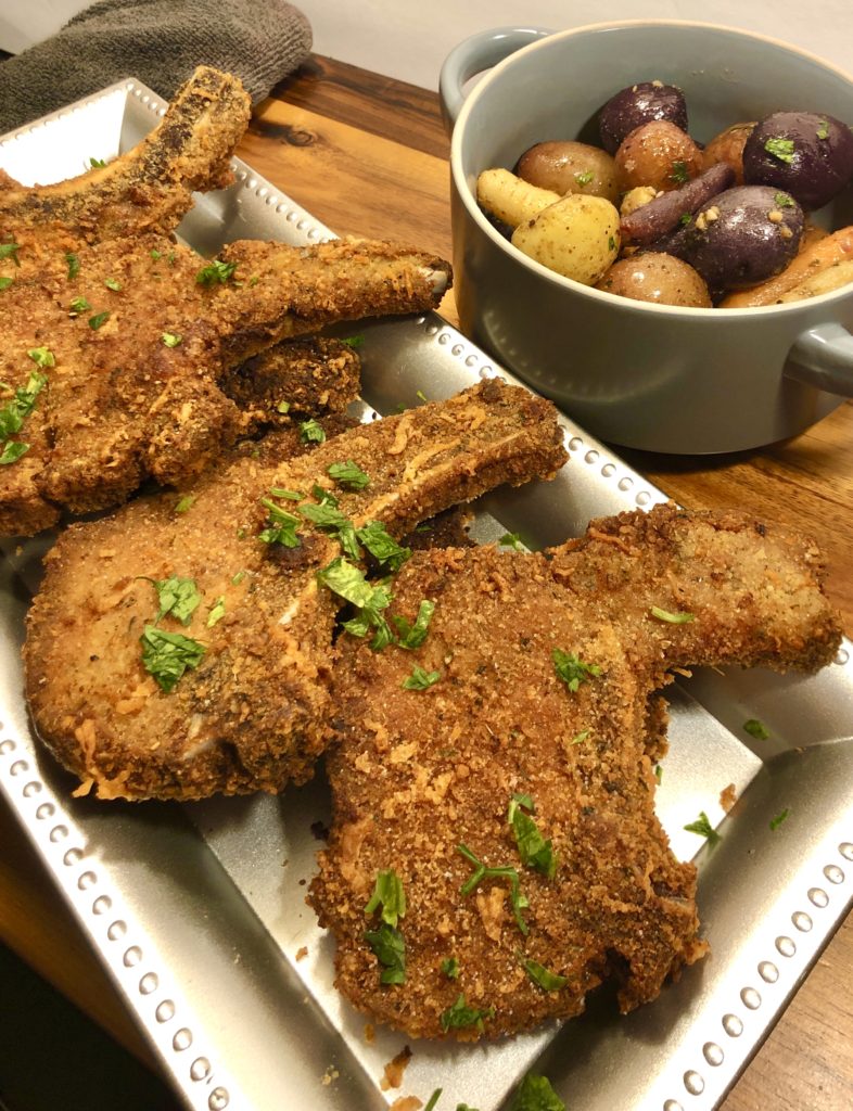 parmesan crusted pork chops on long silver plate on wooden cutting board with gray towel and gray bowl of roasted baby potatoes and rainbow carrots in the background