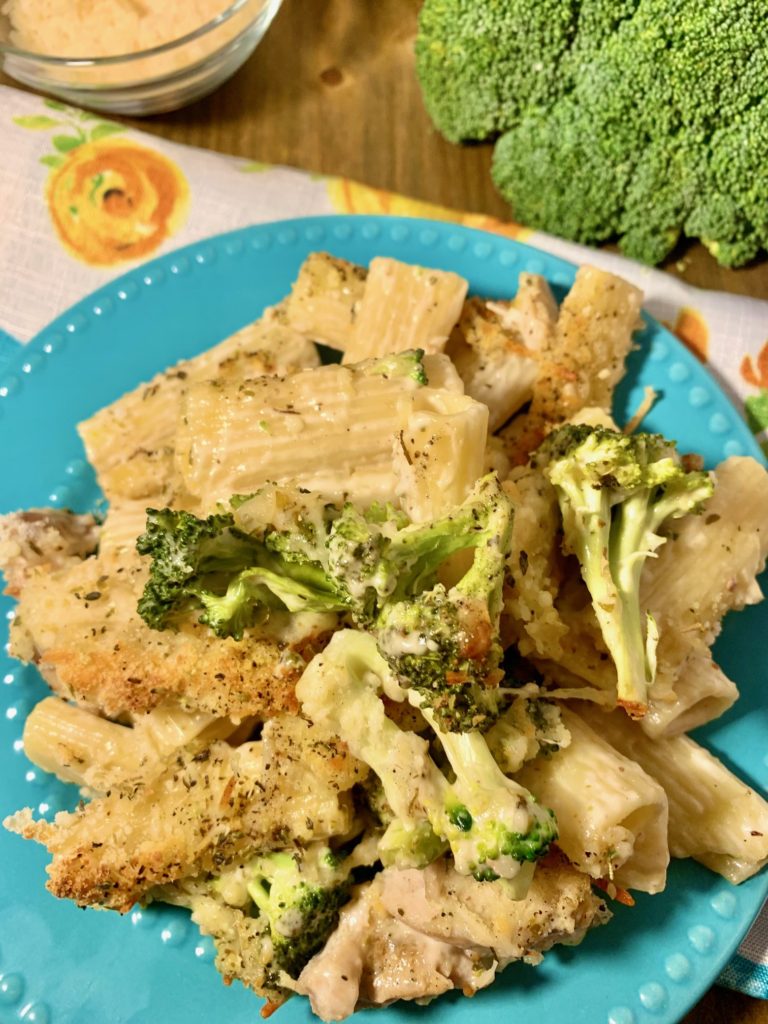 chicken broccoli alfredo ziti with a parmesan cheese and herb topping plated on a light blue plate on top of floral fabric napkin with a head of broccoli and a ramiken of shredded parmesan cheese in the background