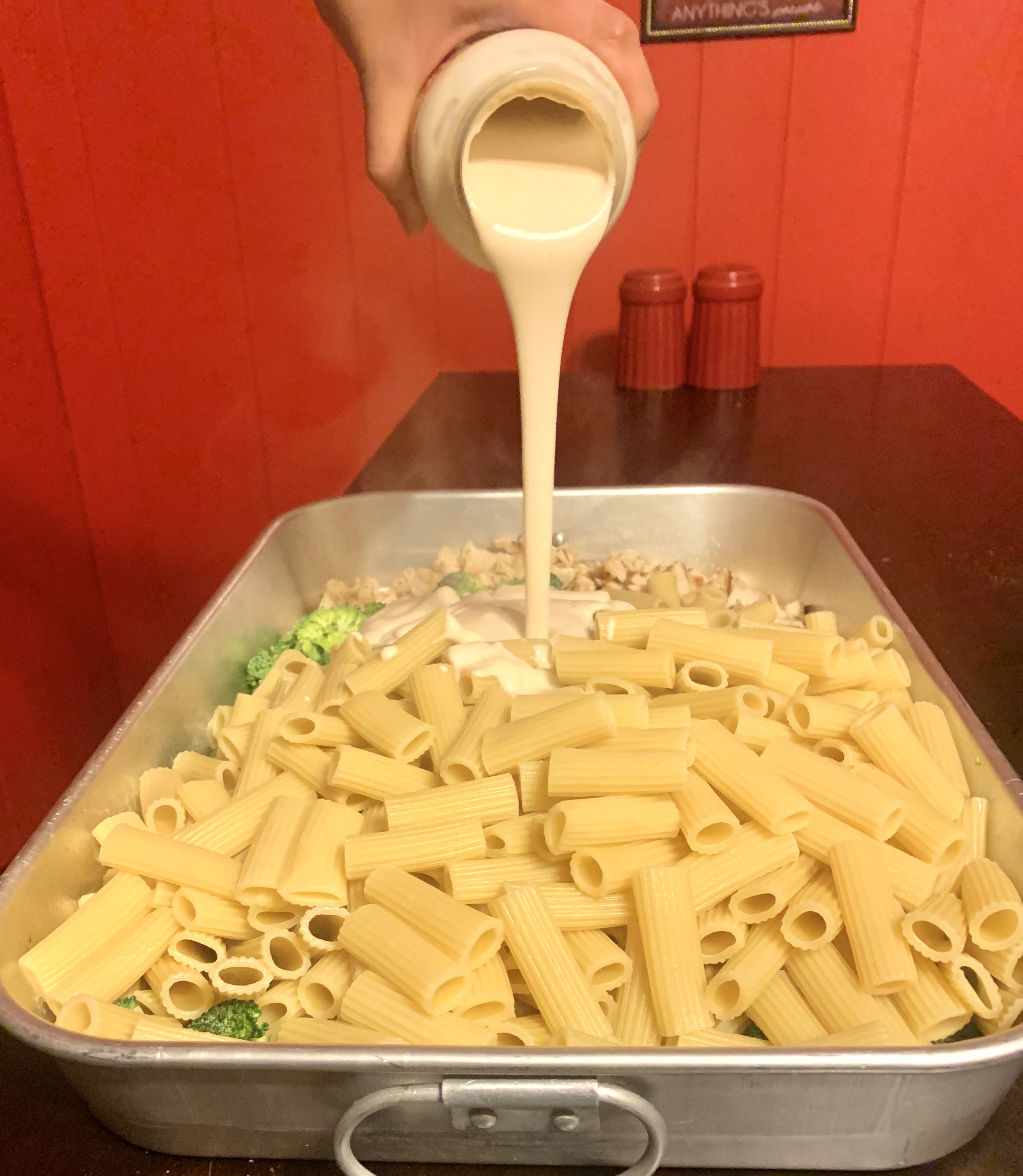 ziti, chicken and broccoli in silver baking pan with jarred alfredo being drizzled over it