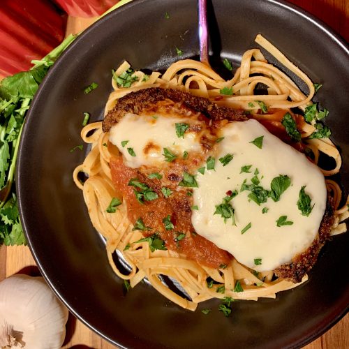 chicken parm main photo with black plate and surrounded by garlic and parsley