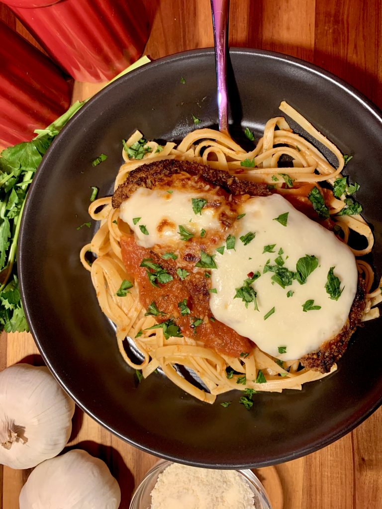 chicken parm main photo with black plate and surrounded by garlic and parsley