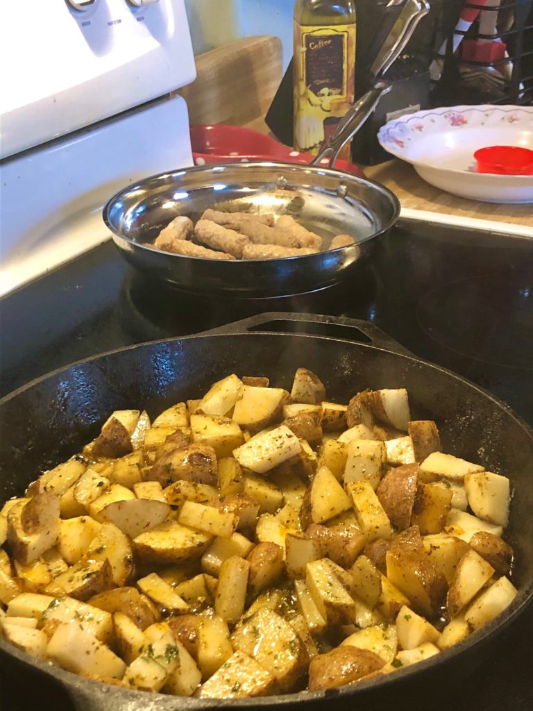 seasoned breakfast potatoes frying in cast iron skillet with small pan of sausage links cooking in the background