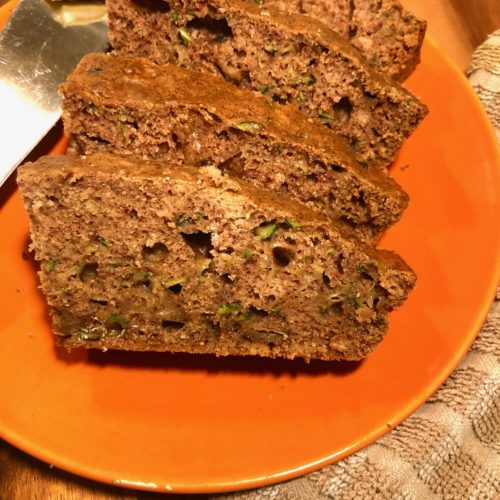 zucchini bread slices plated on orange plate with a pat of butter. the plate is on a wooden cutting board next to a tan and white checkered towel