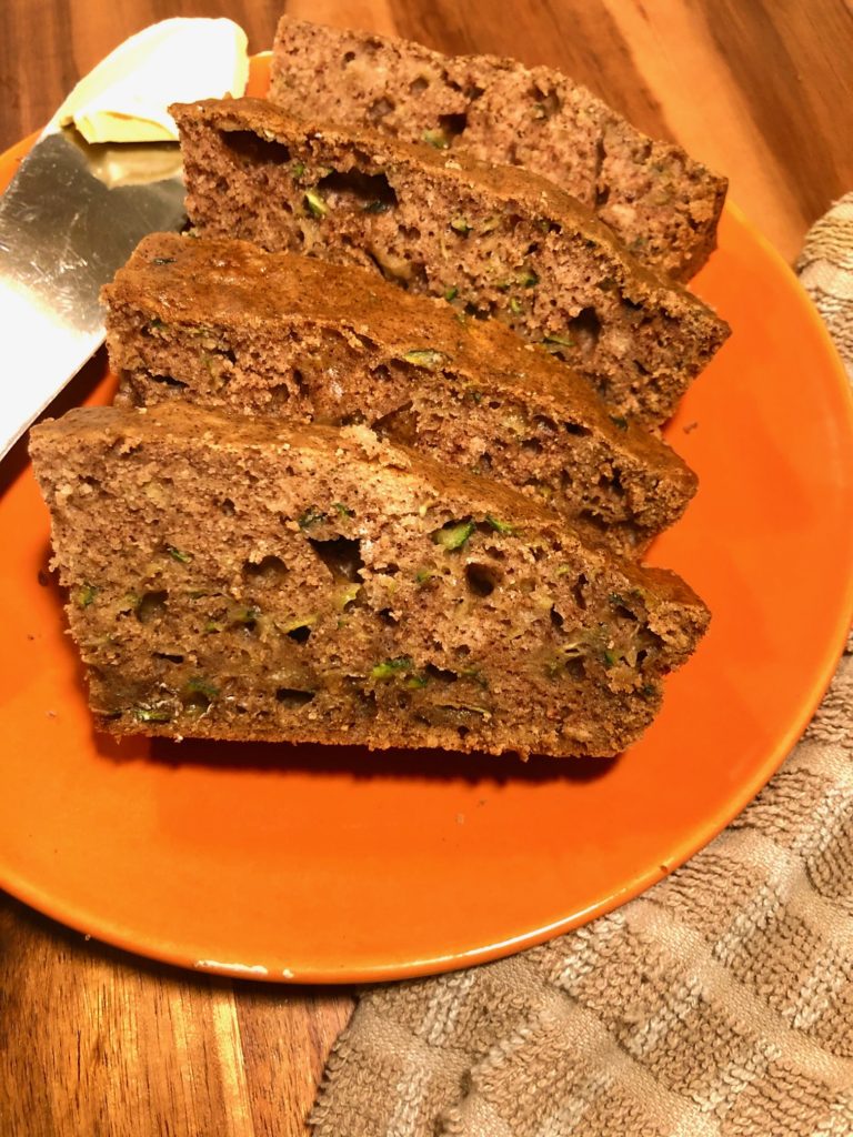 zucchini bread slices plated on orange plate with a pat of butter. the plate is on a wooden cutting board next to a tan and white checkered towel