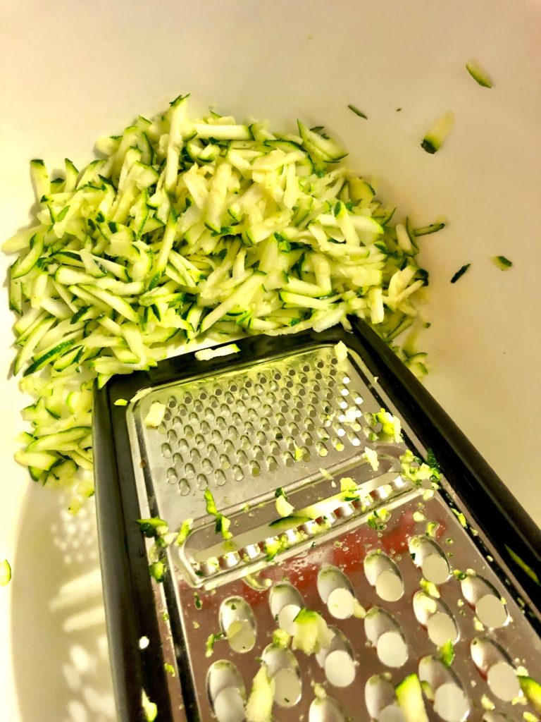 grating zucchini on grater into large white mixing bowl