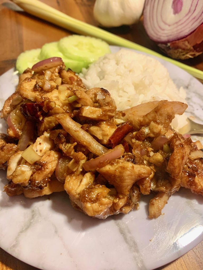 lemongrass chicken with onion and thai chilis. plated on white marble plate with jasmine rice and cucumber slices. plate is on wooden cutting board with purple onion and lemongrass stalk in the background