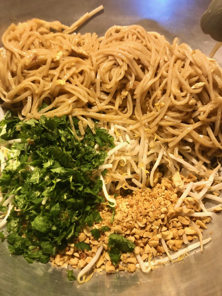 adding stir fried pad thai noodles to large metal mixing bowl with fresh cilantro, bean sprouts and chopped peanuts