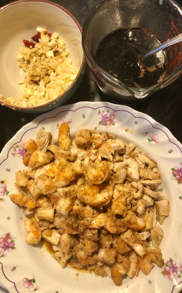 one bowl with garlic ginger and thai chilis. one glass measuring cup with pad thai sauce. a white and floral bowl with cooked chicken tenderloins and shrimp