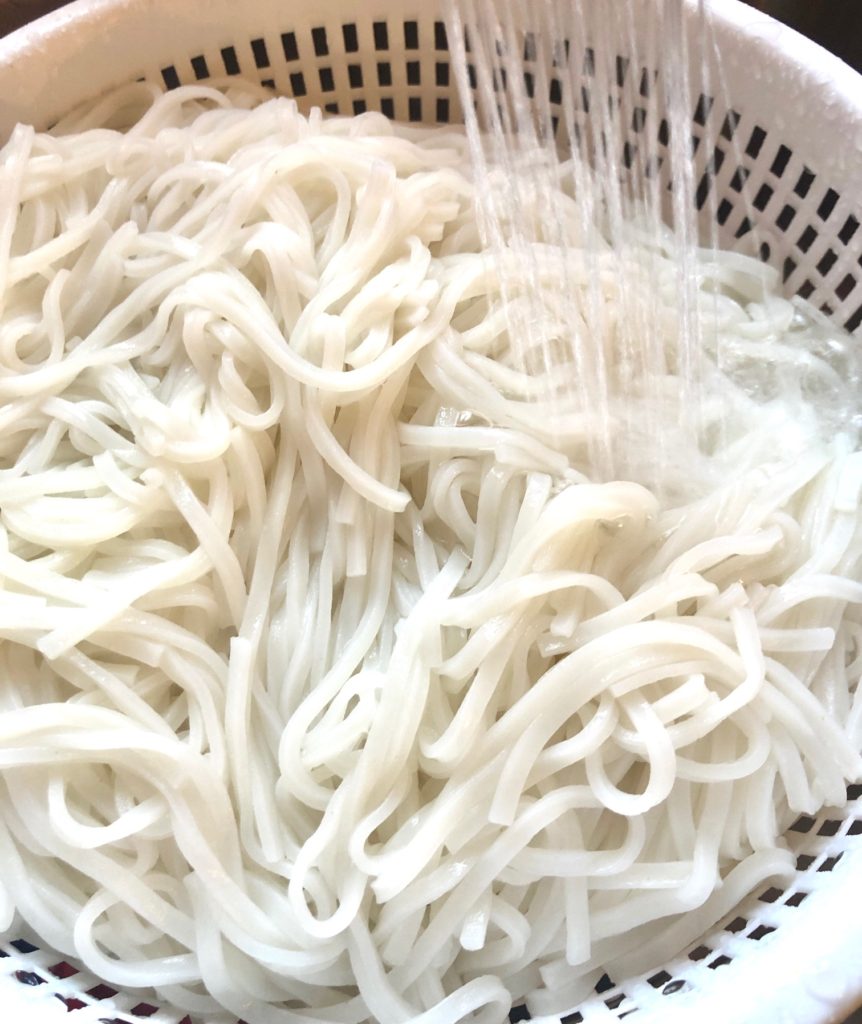 rice noodles being rinsed under cold water in a strainer