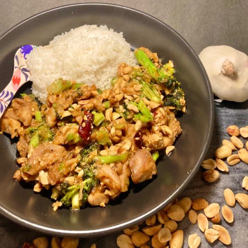 Thai peanut chicken with broccoli served with white rice. Served in black bowl with floral spoon and surrounded on cutting board by whole garlic and peanuts