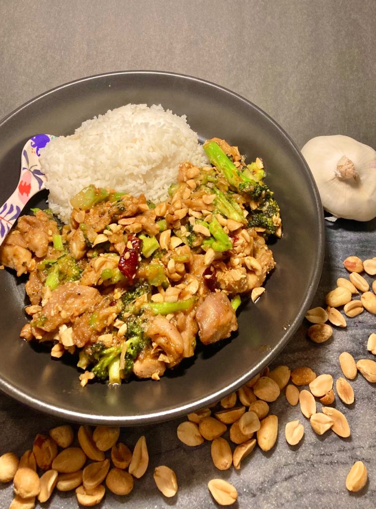 Thai peanut chicken with broccoli served with white rice. Served in black bowl with floral spoon and surrounded on cutting board by whole garlic and peanuts