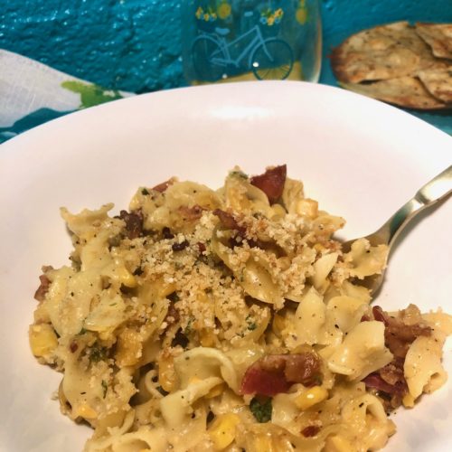 corn chowder casserole with egg noodles and bacon. covered with panko breadcrumb, parm cheese and bacon grease topping. pictured in white square bowl with gold fork. white napkin with blue and yellow flowers. cup with blue bicycle and blue plate with garlic and cheese pita triangles in background