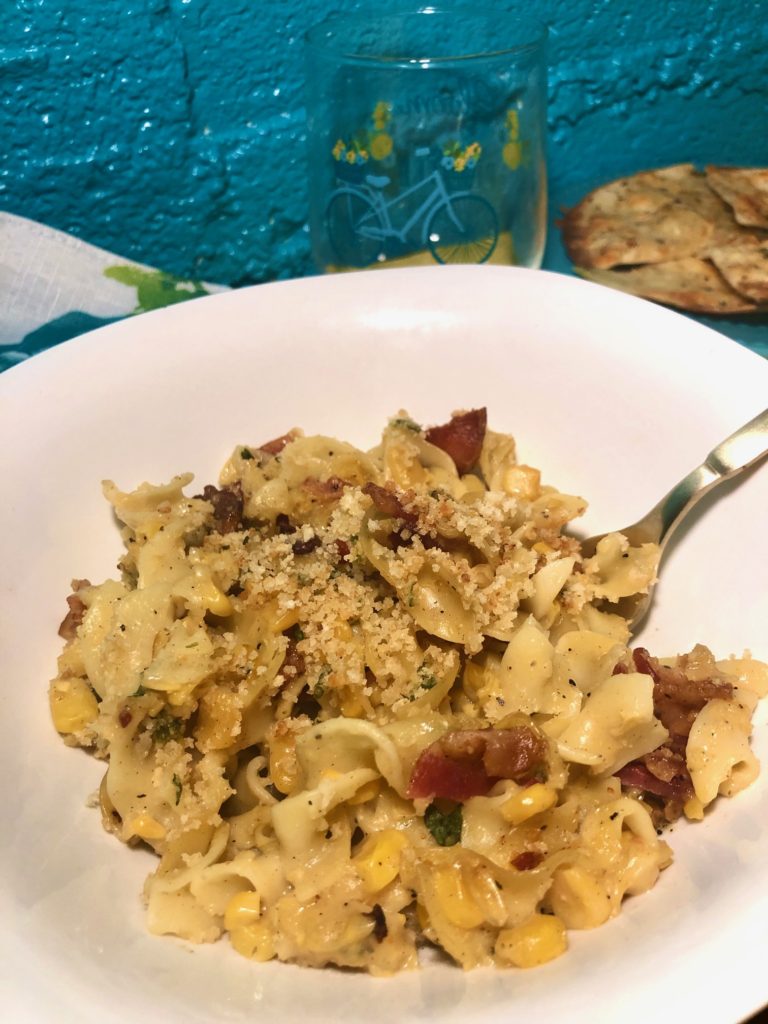 corn chowder casserole with egg noodles and bacon. covered with panko breadcrumb, parm cheese and bacon grease topping. pictured in white square bowl with gold fork. white napkin with blue and yellow flowers. cup with blue bicycle and blue plate with garlic and cheese pita triangles in background