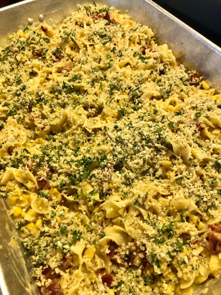 egg noodles mixed with corn chowder and bacon and topped with a panko bread, parsley, parmesan cheese and bacon grease topping. pictured in silver casserole dish before baking