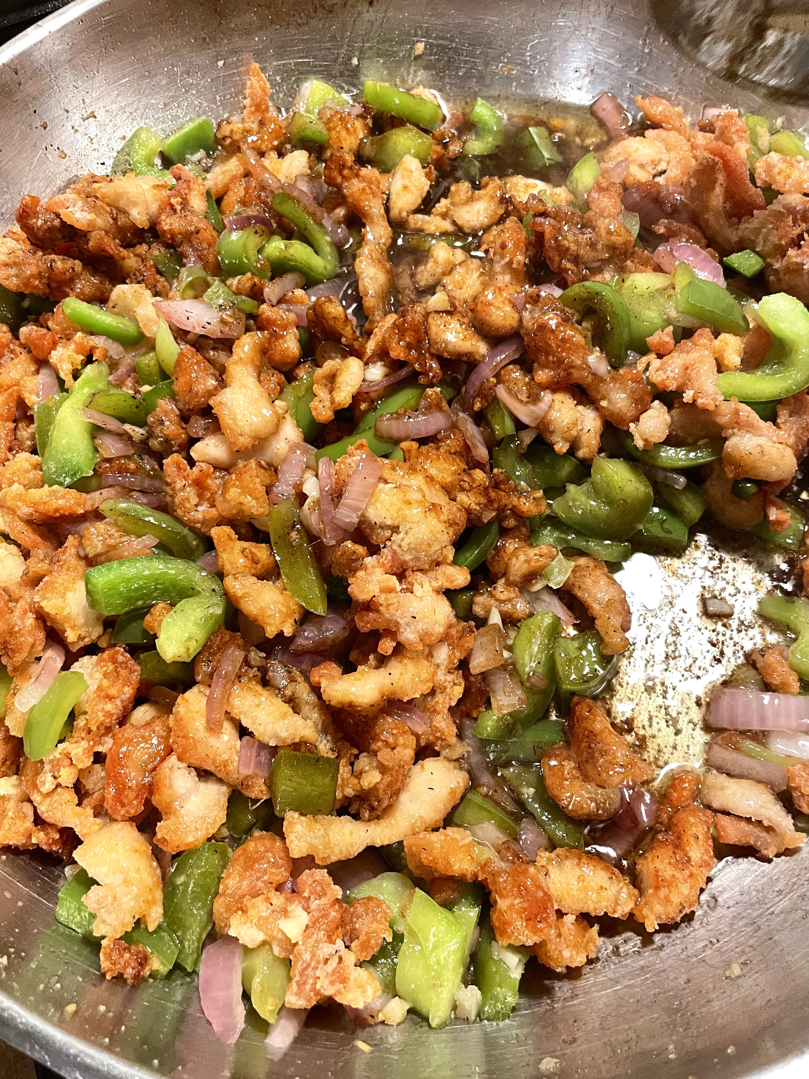 Stirring Pepper Chicken in a large pan. The pan has garlic, green peppers, onion, fried chicken and a pepper sauce.
