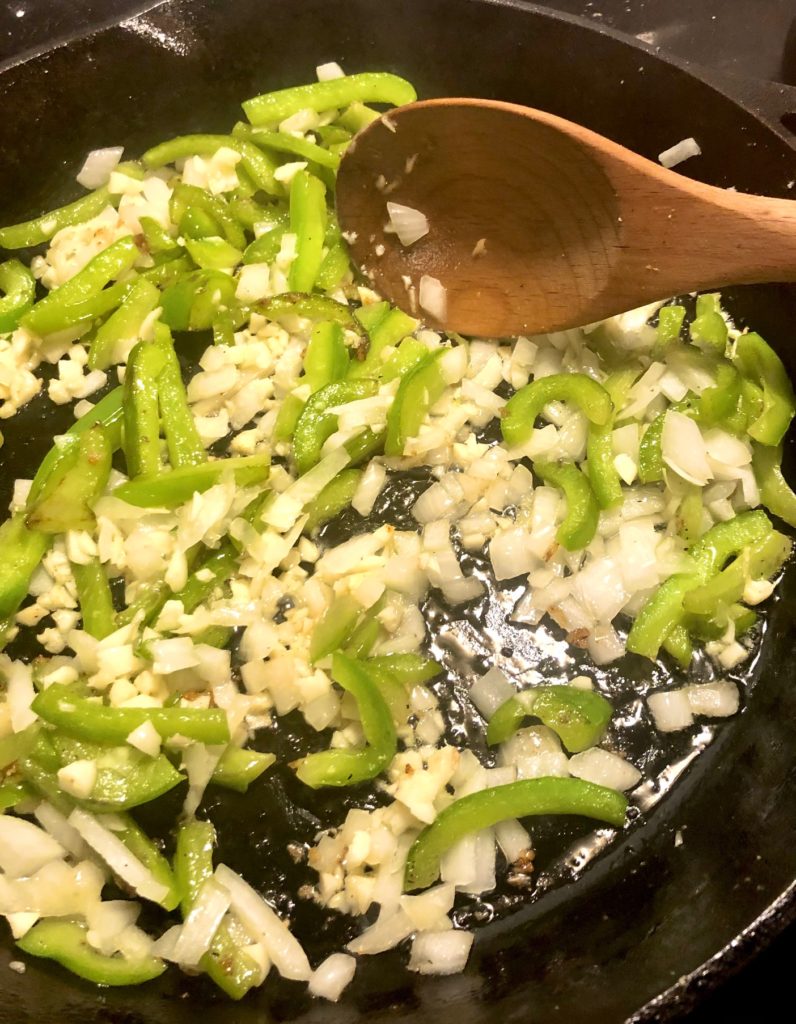 green peppers, onion and garlic cooking in black cast iron skillet and being stirred with wooden spoon