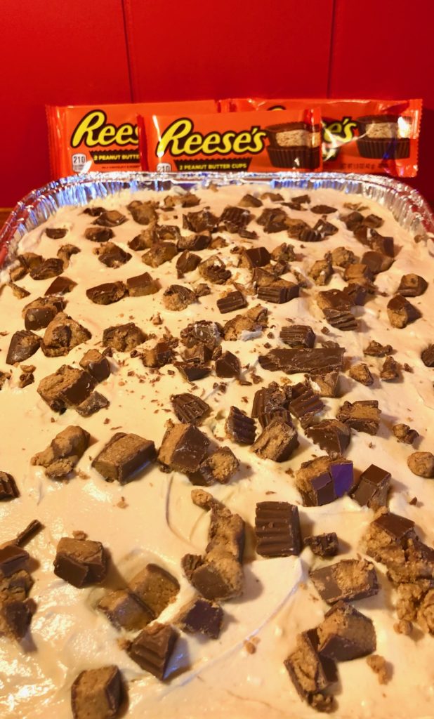 dark chocolate cake with peanut butter and cream cheese whipped frosting and topped with chopped Reese's cups. Pictured in a foil pan in front of red wall with three packs of Reese's cups propped up against it.