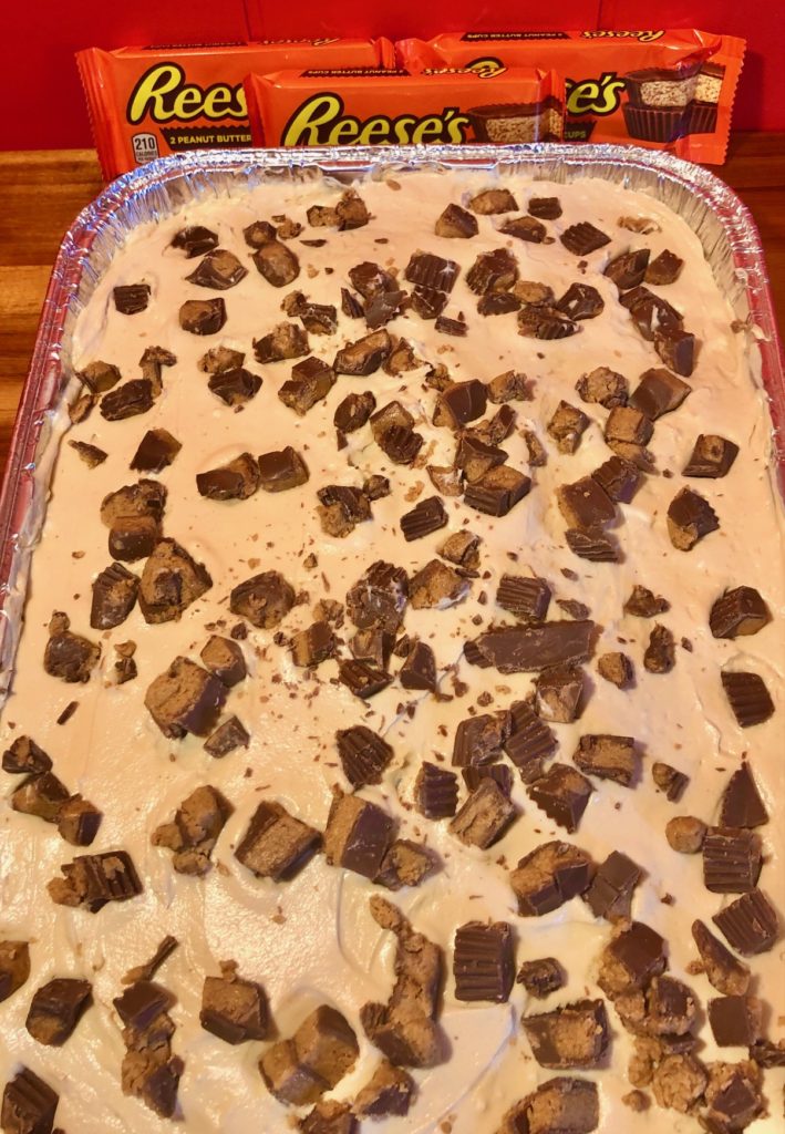 chopped reese's peanut butter cups on top of a peanut butter frosting frosted cake in a foil pan. pictured in front of packages of Reese's cups