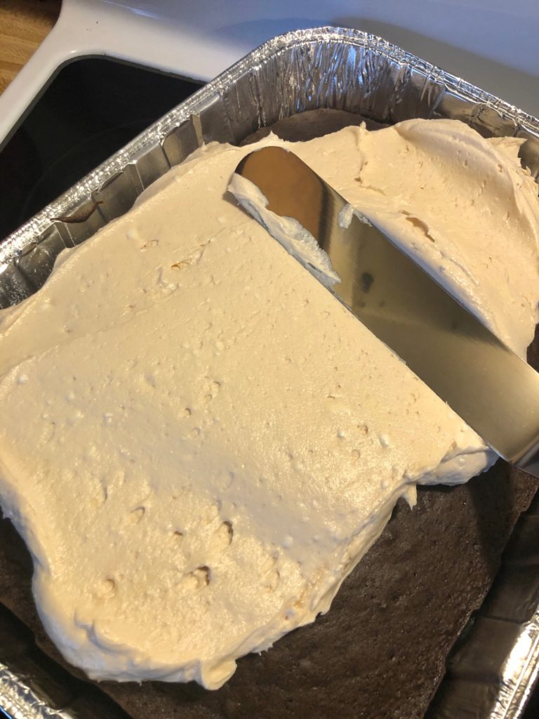 dark chocolate cake in foil dish. off set spatula is spreading peanut butter cream cheese frosting