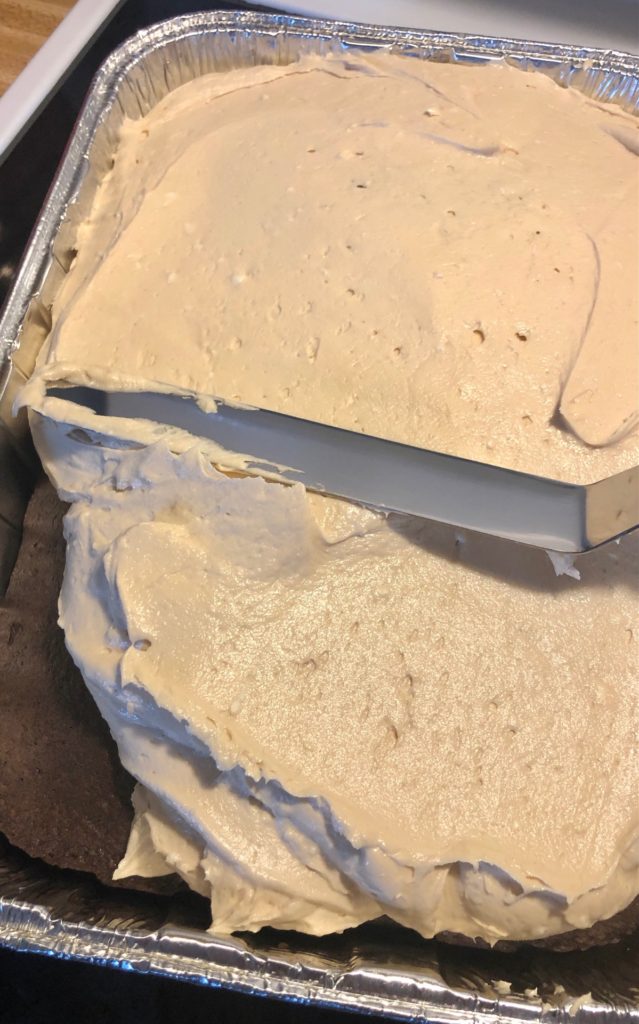 dark chocolate cake in foil dish. off set spatula is spreading peanut butter cream cheese frosting