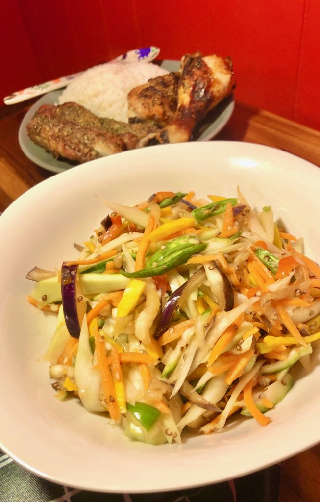 thai sum tom plated in white square bowl with a gray plate of jasmine rice, chicken drumsticks and ribs in the background. The gray plate has a floral spoon with the food
