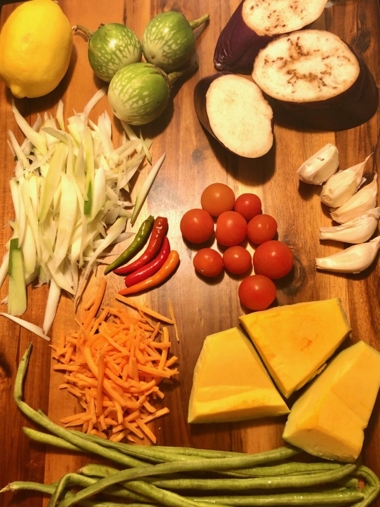 som tum ingredients pictured prior to prepping on wooden cutting board with eggplant, thai eggplant, lemon, papaya, thai chilis, cherry tomatoes, cloves of garlic, shredded carrots, buttercup squash, green beans.
