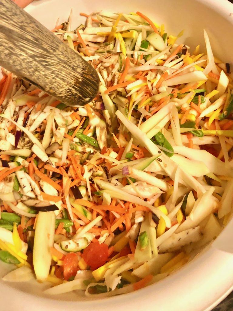 thai som tum vegetables are cut up in a large white mixing bowl and being pounded by a wooden pestle