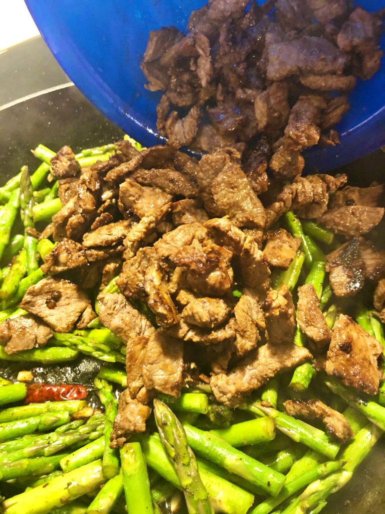 asparagus and garlic frying in black skillet and cooked flank steak being added to pan from blue bowl