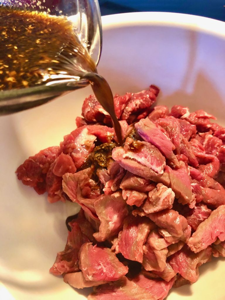 thinly sliced flank steak in white mixing bowl with marinade being poured in