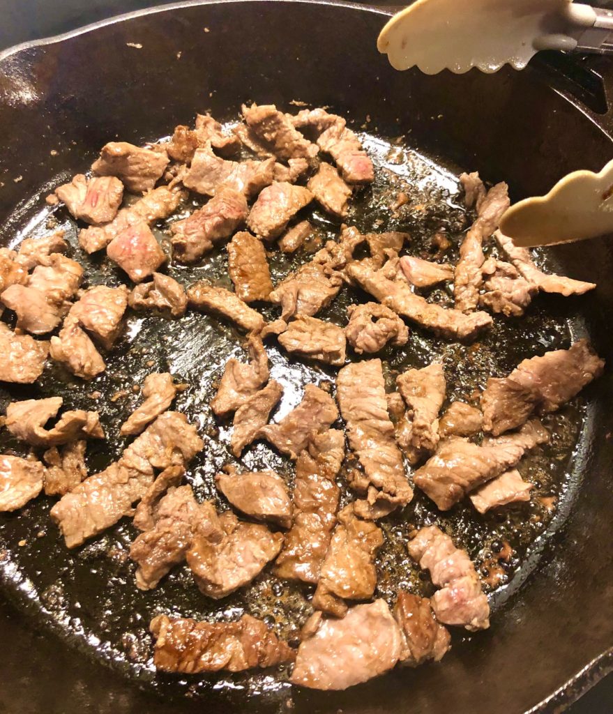 flank steak frying in black skillet pictured with tongs