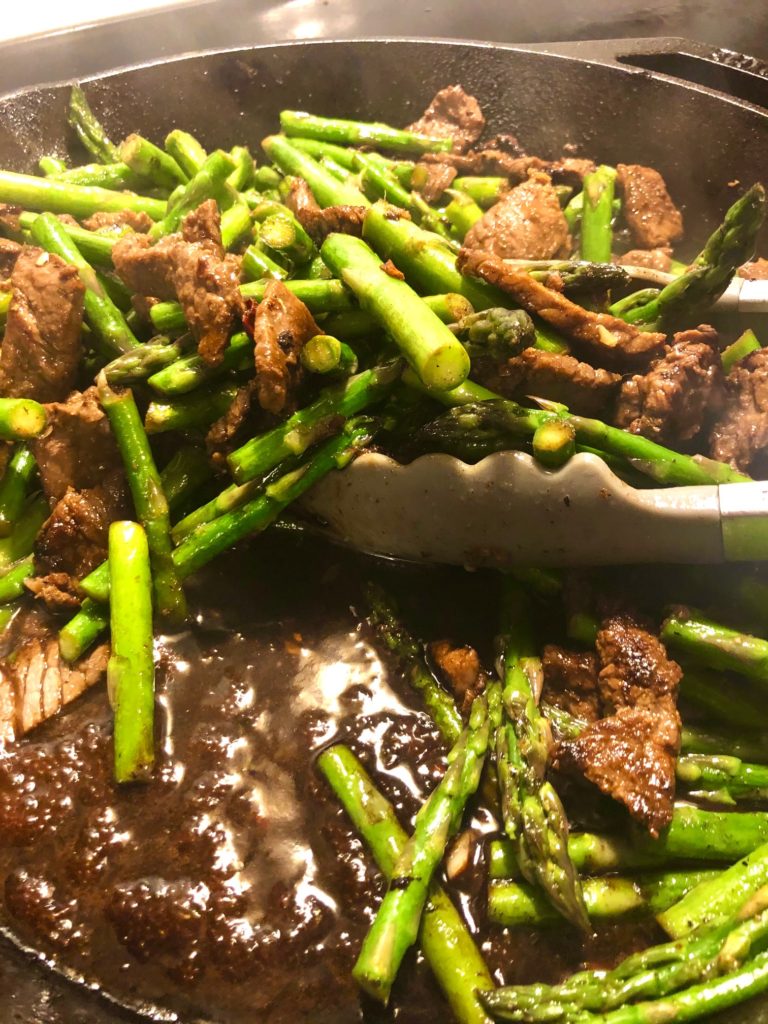 steak and asparagus stir-fry in cast iron skillet being mixed with silver tongs