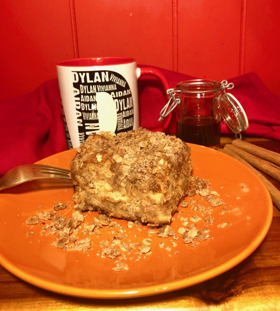 french toast casserole on orange plate surrounded by oats. pictured with a gold fork, a white coffee mug, jar of syrup and cinnamon sticks on a wooden table