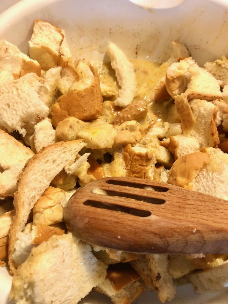leftover day old bread being mixing with french toast egg mixture with wooden spoon in large white mixing bowl
