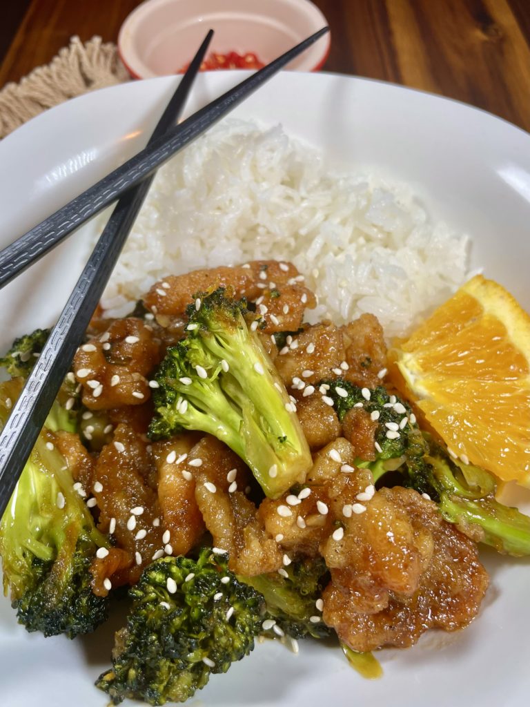 Orange Chicken with Broccoli pictures on a white square plate with Jasmine rice and a slice of orange. Black chopsticks are laying across the plate. There is a ramekin with fresh Thai chilis in the background.