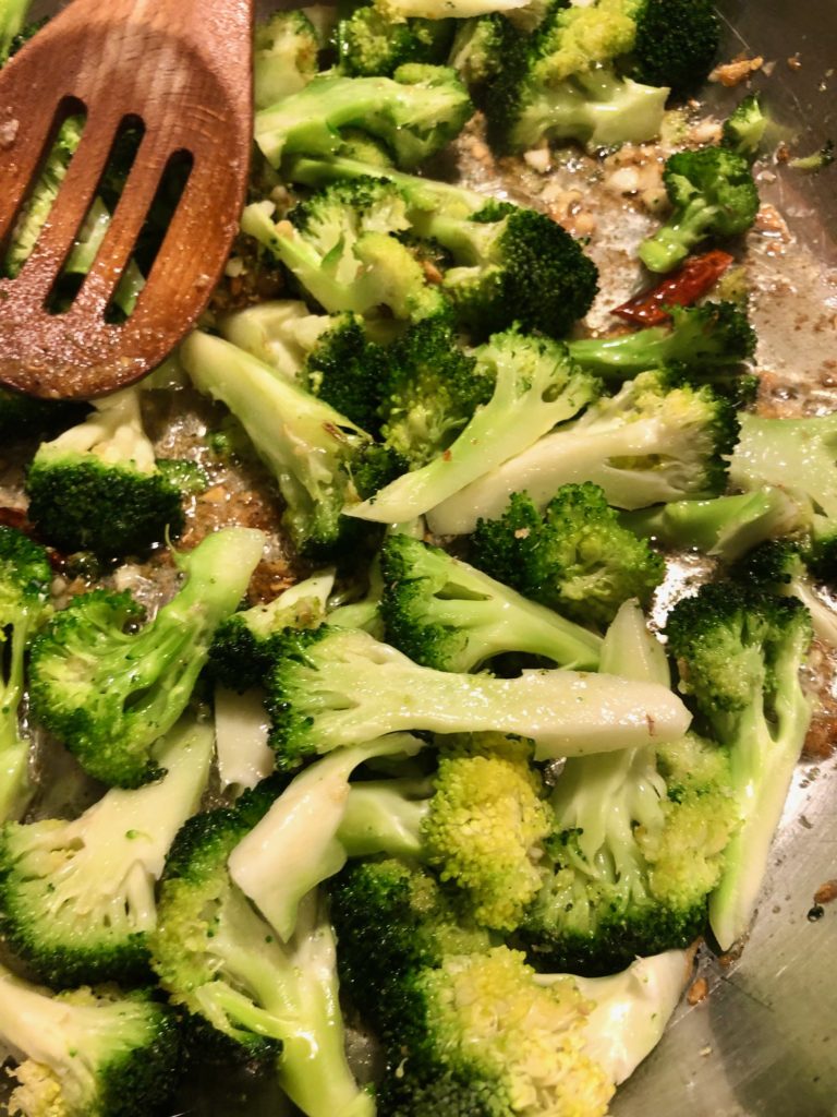 broccoli, Thai chilis, garlic and ginger frying in large silver frying pan and being stirred with wooden spoon