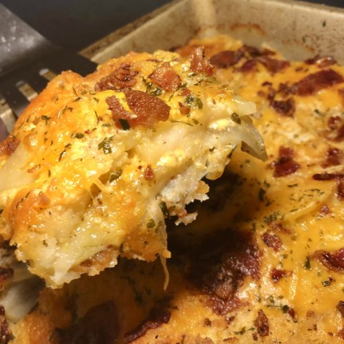 creamy potatoes au gratin with bacon. a corner piece is being held up out of the baking dish on a spatula so that you can see the layers of potato, onion and cheese