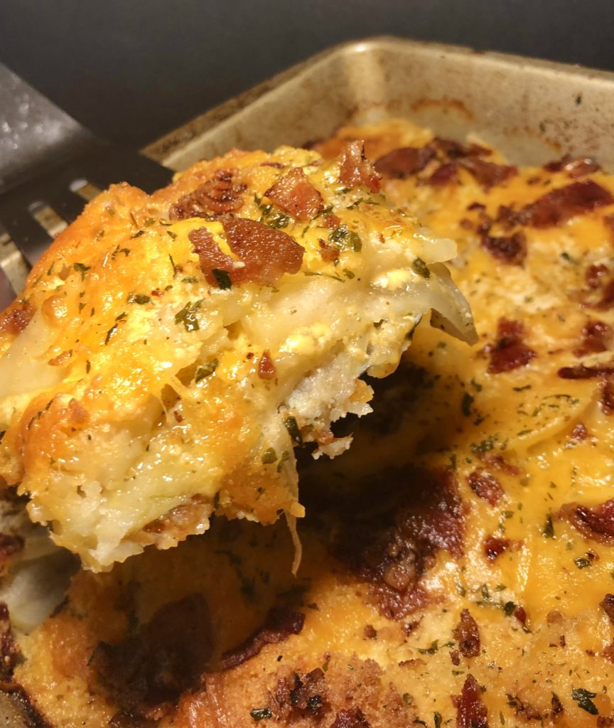 creamy potatoes au gratin with bacon. a corner piece is being held up out of the baking dish on a spatula so that you can see the layers of potato, onion and cheese