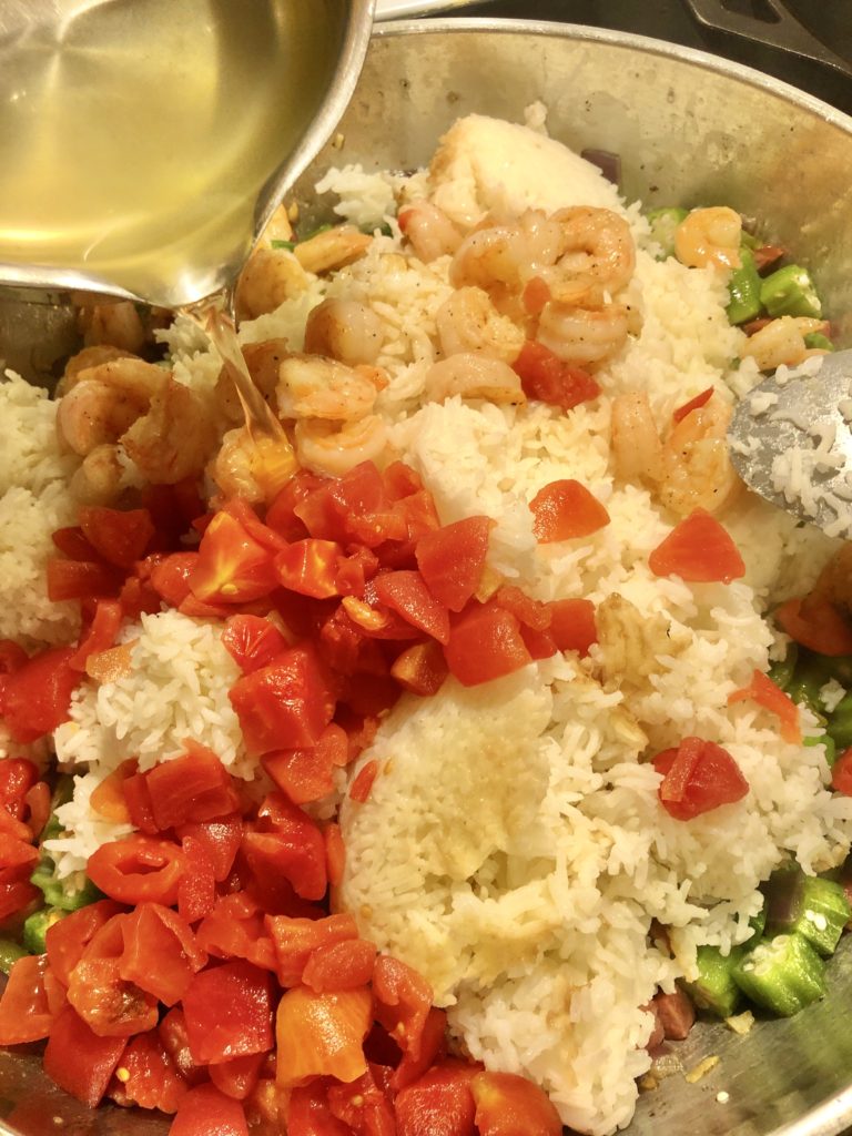 large skillet with rice, green pepper, diced tomatoes, shrimp and kielbasa with chicken broth being poured in